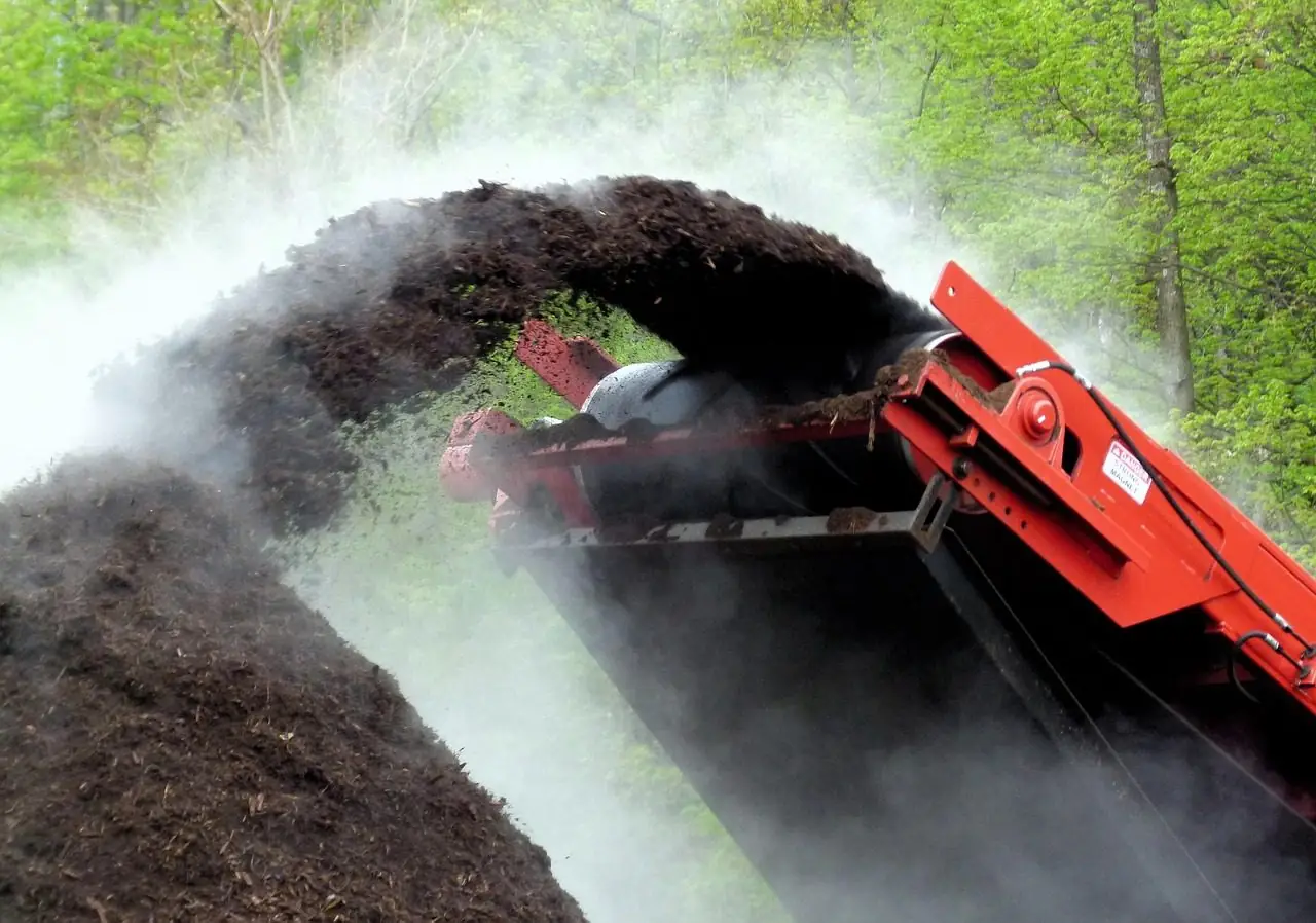 Red conveyor throwing a stream of dark ground mulch onto a steaming pile
